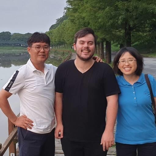 three people pose for a picture, standing in front of a lake. They are all smiling.