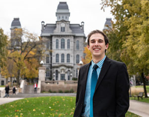 a man stands in front of a gothic building. He is smiling and wearing a suit.