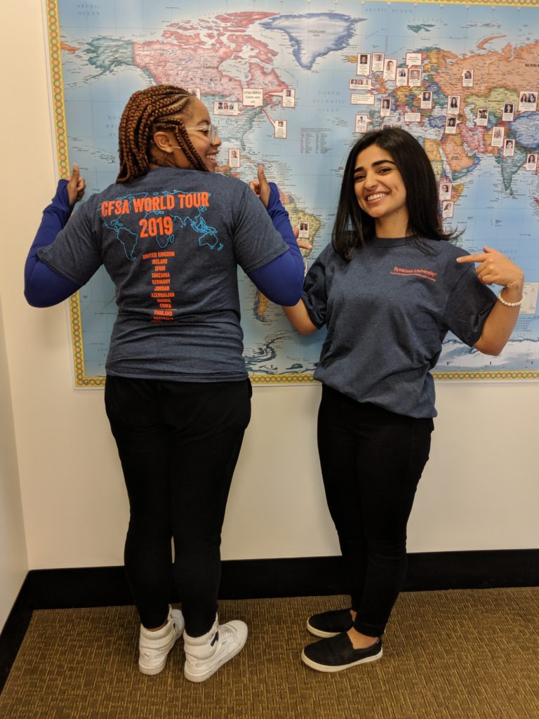 two women stand in front of a picture of a map. One has her back to the camera, and one her front. Both are smiling and giving thumb's up. They are wearing new CFSA blue tshirts.