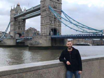Zach Watson stands leaning on a wall in front of the Thames River in London, England. The London Bridge can be seen in the background, as well as a cloudy, grey sky.