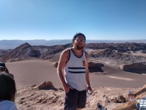 white man standing in a desert and smiling. He is wearing an SU tanktop