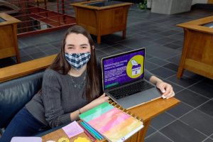 Woman in mask sits in front of computer and notebooks