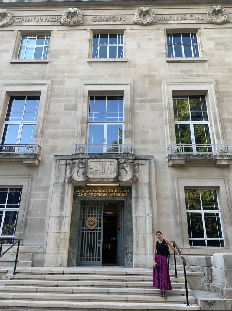 Woman stands in front of grey stone building that reads "London School of Hygiene and Tropical Medicine"