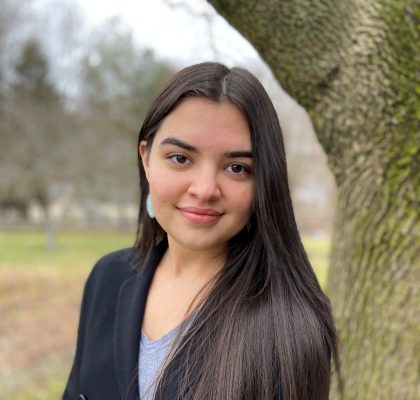 headshot of a fair woman with long, dark brown hair smiling into the camera as she leans next to a tree