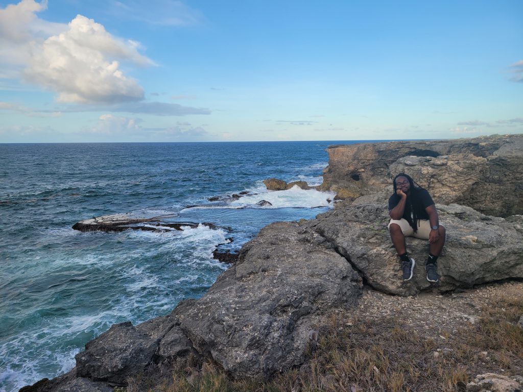 a black man with long hair sits contemplatively facing the camera on a rocky cliff to the right of a beautiful ocean view