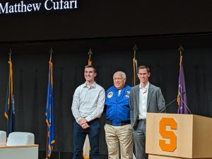 Alex Metcalf, Col. Fred Gregory, and Matt Cufari stand in a line smiling for photos on the stage of the NVRC, standing in front of a row of military flags and a podium with a large orange block S
