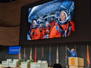 Older man in blue jacket (Col. Gregory) stands behind a podium on a stage pointing to a large powerpoint slide which features an image of him as a young man with his spaceflight crew on a Shuttle mission.