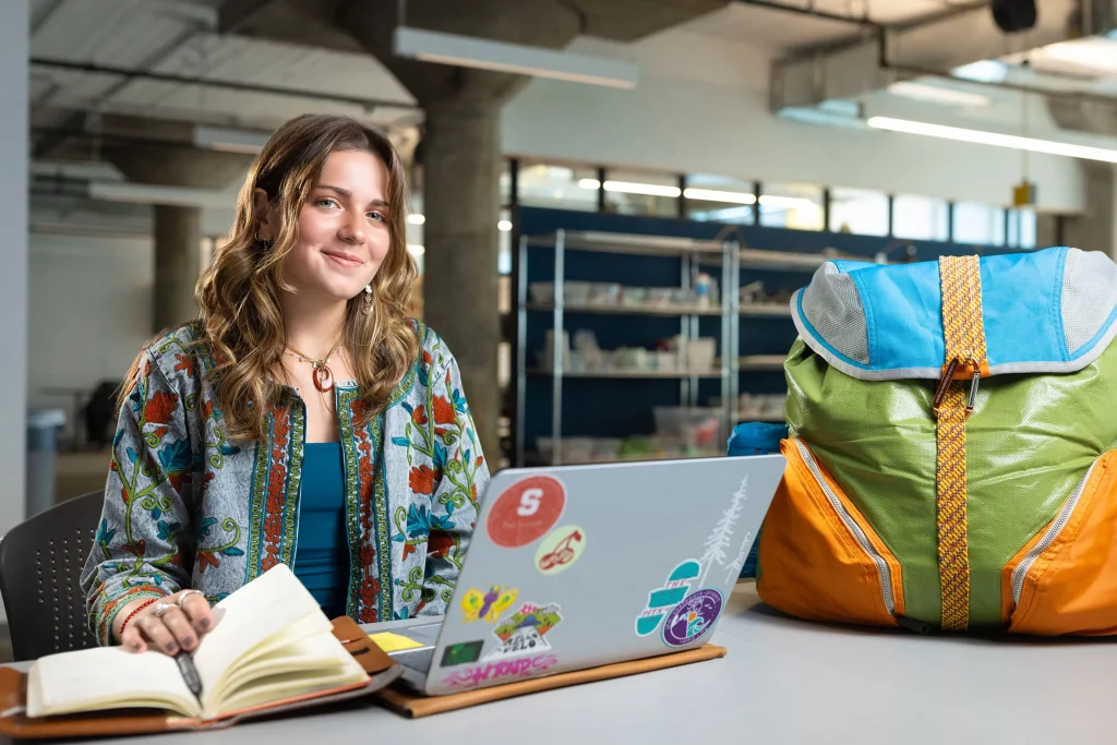 Lilyan sits behind a desk with her computer and a book in front and the backpack she made to the right. Her laptop is decorated in colorful stickers and the backpack is made of blue, green, orange and gray repurposed outdoor gear.