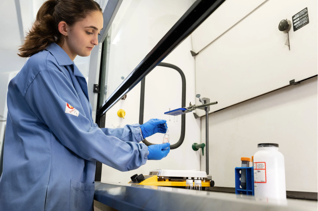 Woman stands above a sink in a scientific lab, looking down at her hands holding equipment. She wears a blue lab coat and blue gloves.