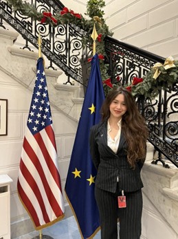 Mikah stands smiling in a business suit in front of the EU and U.S. flags near a stairwell decorated for the winter holidays.