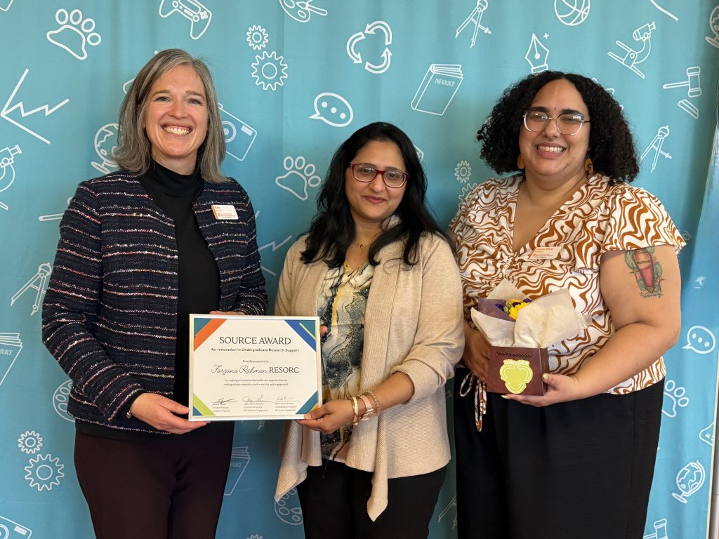 Kate Hanson, Farzana Rahman, and Odette Rodriguez stand smiling in front of a blue backdrop. Two hold onto the corners of a framed certificate and one holds a container of flowers.