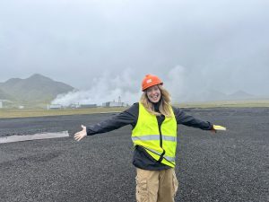 A woman with blonde hair stands smiling in a yellow construction vest and orange hard hat with her arms spread out. She stands in front of a foggy Icelandic geothermal research site.